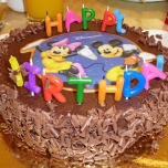 Geburtstagstorte aus Schokolade mit einem Rand aus Schokostreuseln, in der Mitte ein Bild von Mickey mouse und Minnie Mouse. Buchstaben Kerzen bilden das Wort Happy Birthday