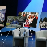 Bühne mit Sesseln und einem kleinen Tisch, dahinter Fotos auf Ständern