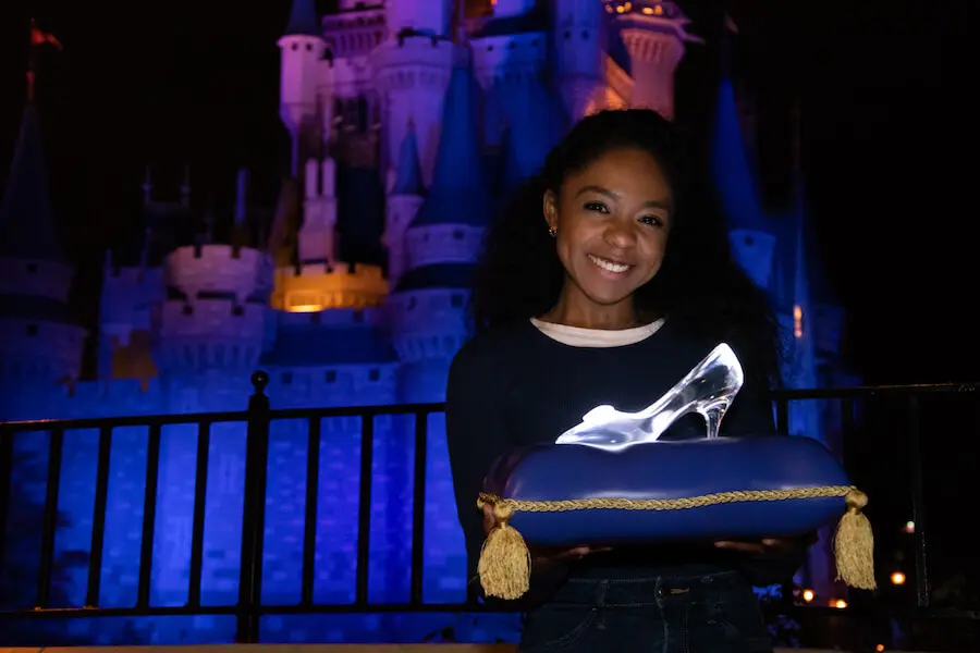 Foto mit Cinderellas Slipper eine junge dunkelhäutige Frau hält ein Kissen mit Cinderellas Glasschuh vor dem Cinderell Castle in den Händen und lächelt in die Kamera