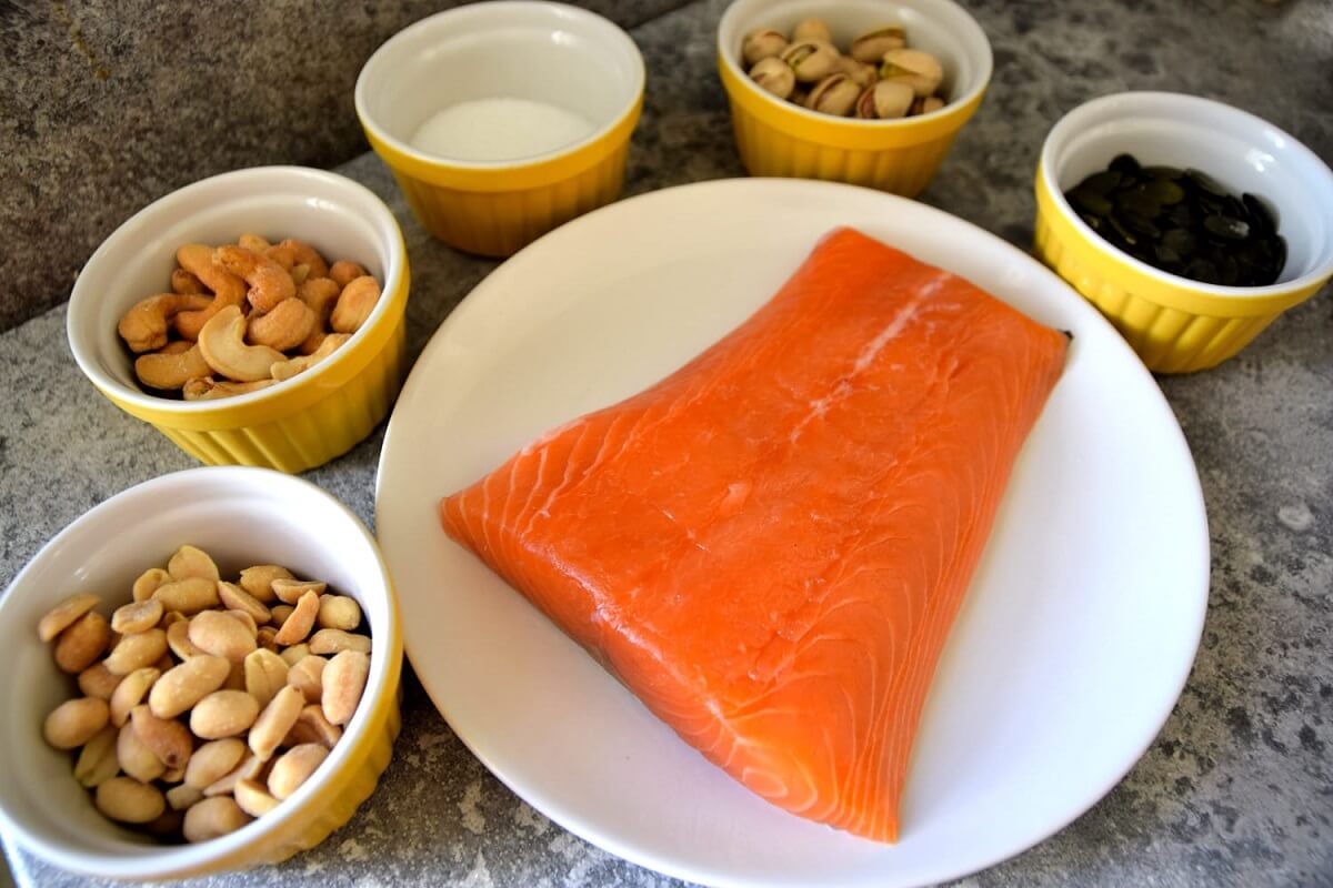 Zutaten für Nut Crusted Salmon Auf einem Teller liegt ein Stück Lachsfilet, darum herum stehen mehrere Schälchen mit verschiedenen Nüssen und etwas Zucker