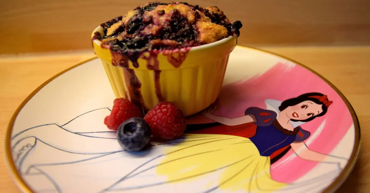 Berry Cobbler auf Schneewittchenteller Auf einem Teller mit Schneewittchen-Motiv steht ein Schälchen mit leckerem Berry Cobbler mit einigen frischen Him- und Blaubeeren