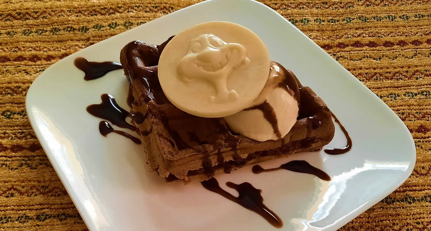 Leckere Schokoladenwaffel Auf einem kleinen quadratischen weißen Teller liegt eine Schokoladenwaffel mit gefrorener Espresso-Mousse und einem weißem Schokoladentaler mit einer Abbildung von Timon aus Disney's König der Löwen