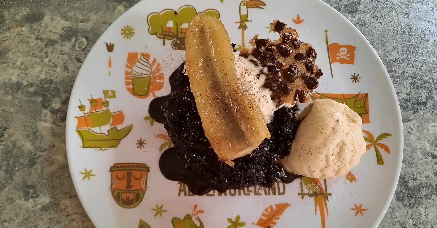 Kungaloosh mit Banae und Eis Ein Schokoküchlein mit Schokoladensauce, karamellisierter Banane, Vanilleeis, Sahne und Cacao Nib Tuile auf einem Teller mit Adventurelandmotiv
