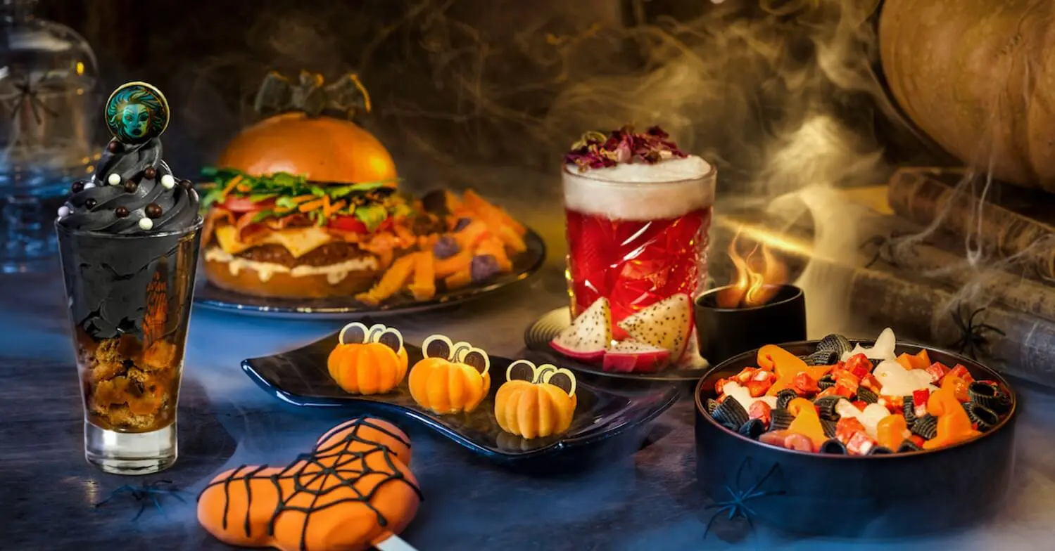 Halloween Gerichte Snacks und Getränke im Halloweenlook sind zur Präsentation angerichtet. Orangene Kürbisküchlein in der mitte des Bildes, davor ein großer Mickey-Mouse Lolli mit Spinnennetz, ein rot dampfender Cocktail, ein Burger und andere orangefarbene Speisen