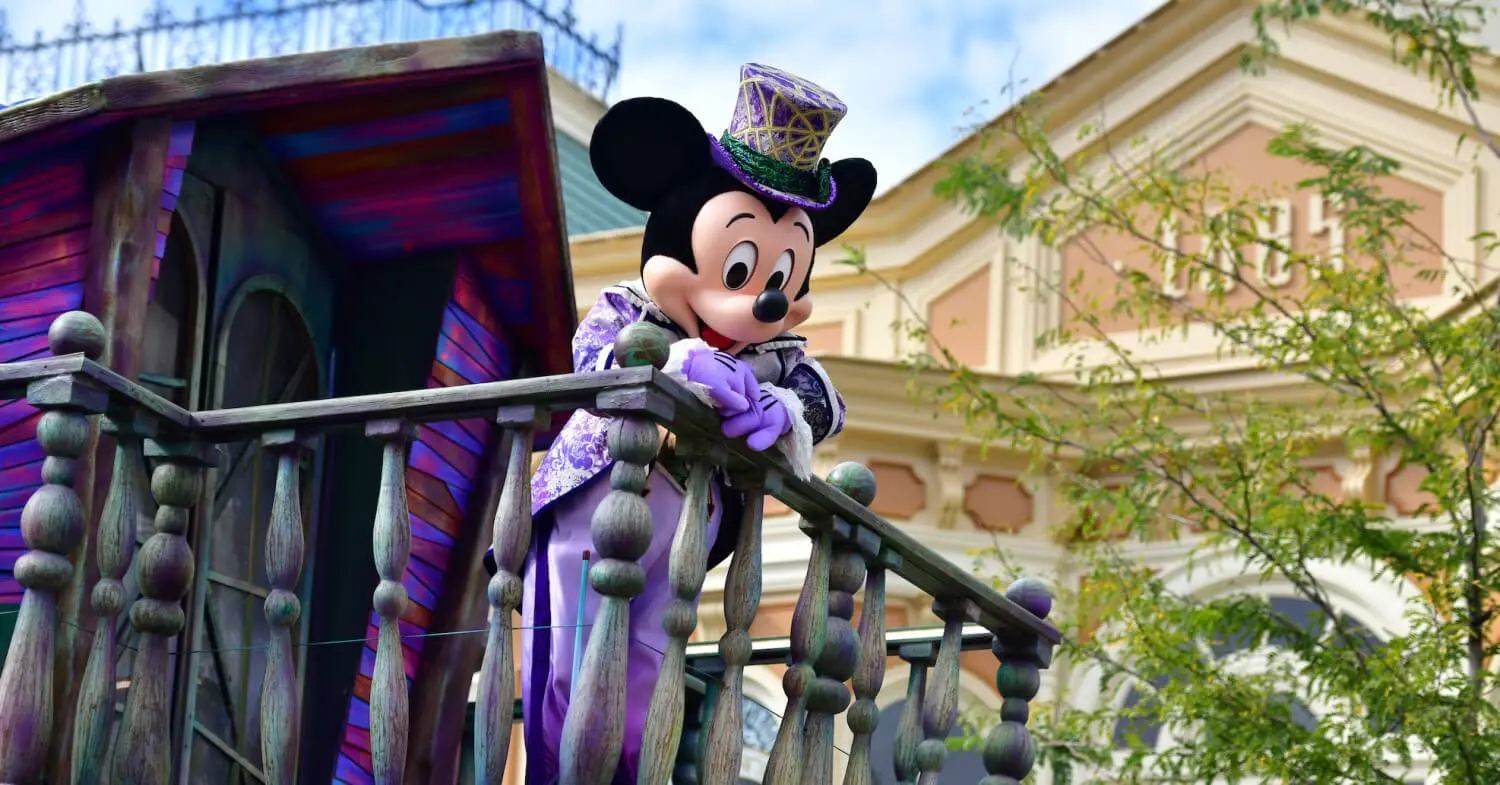 Halloween Mickey Mouse Mickey Mouse in lilafarbenem Frack mit Zylinder, lehnt an einem hölzernen Geländer vor einem Holzhaus
