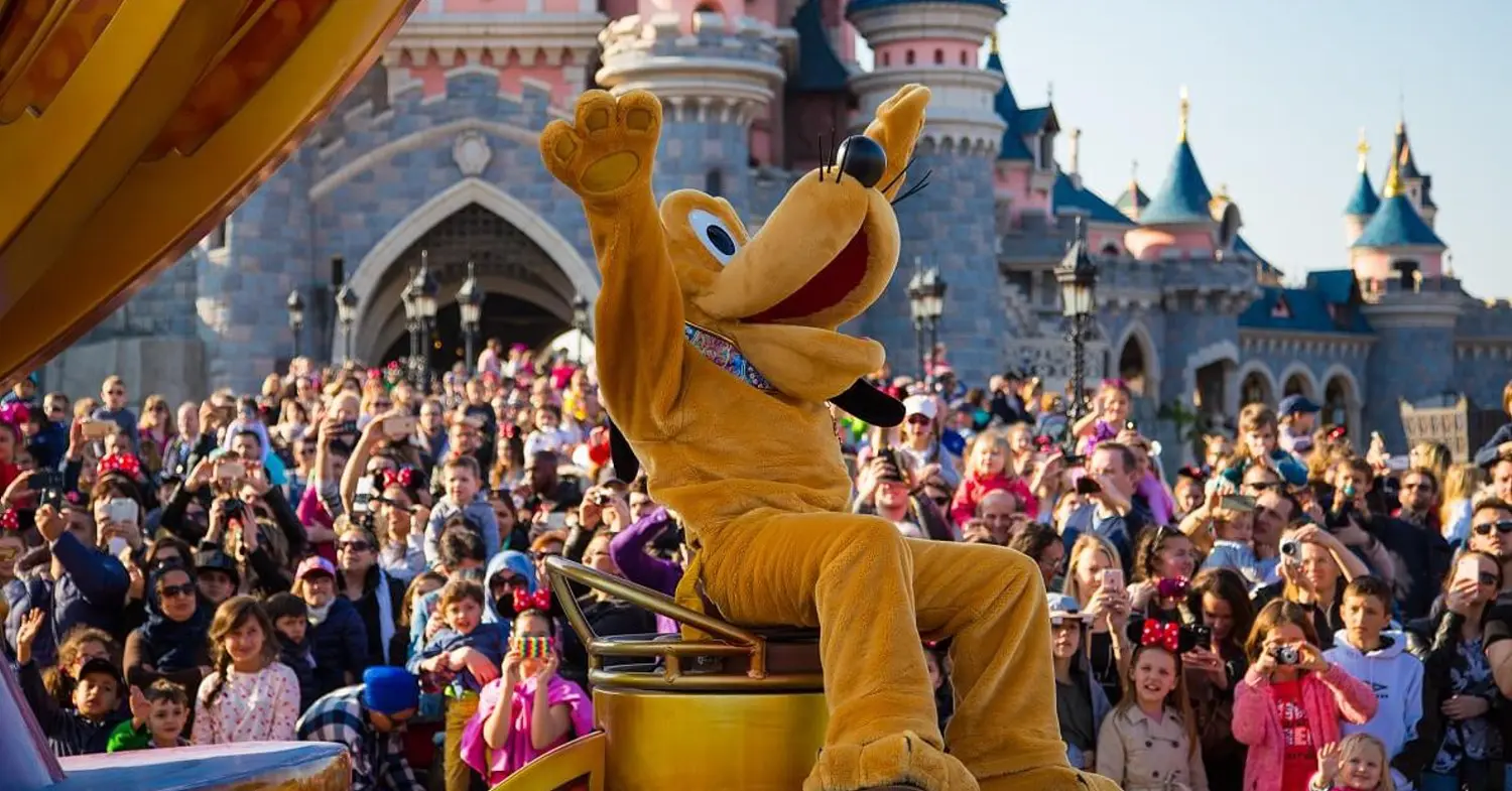Parade mit Pluto Pluto sitzt auf einem Paradenwagen, der gerade am Sleeping Bauty Castle vorbeifährt. Menschenmassen schauen ihm zu.