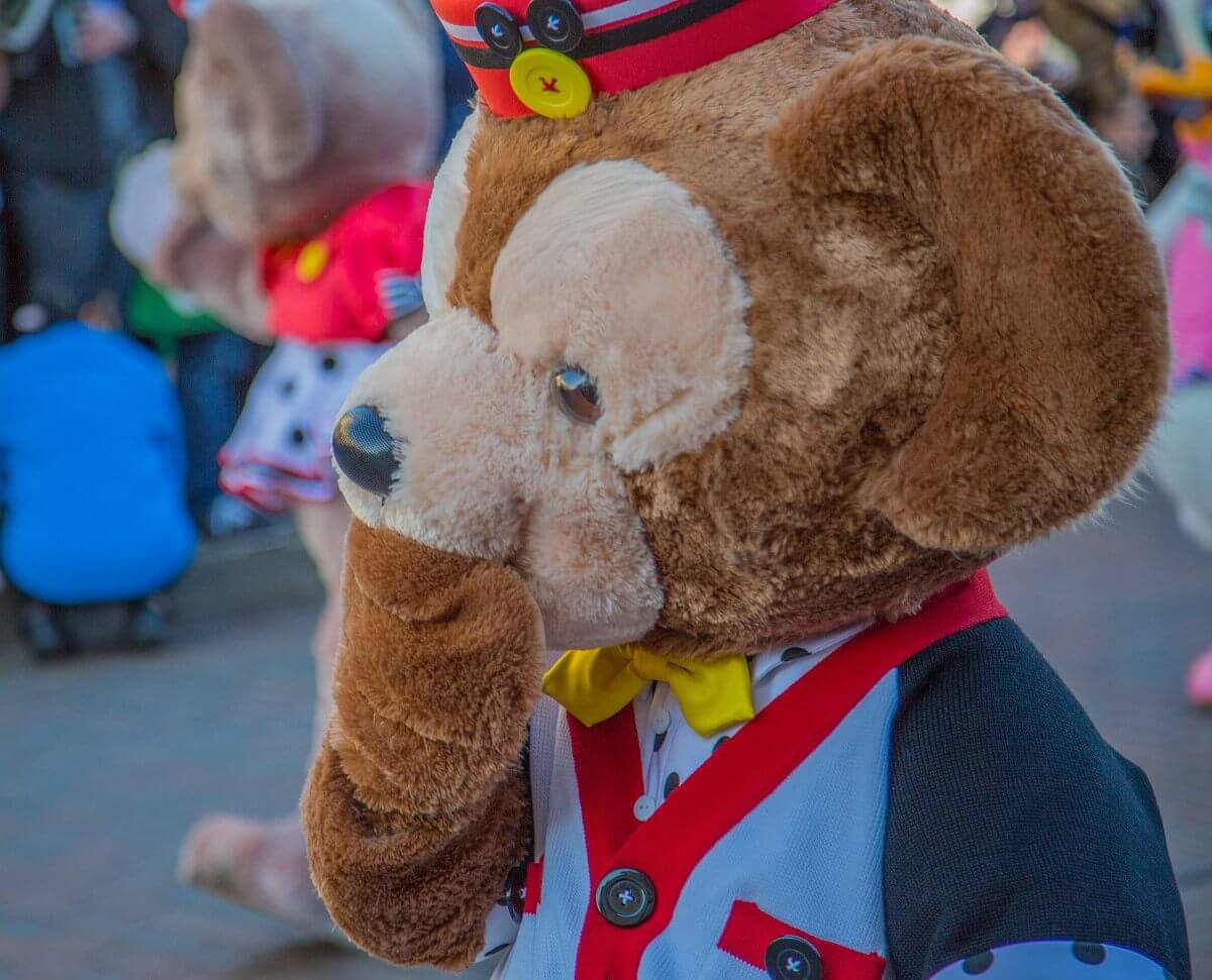 Duffy der Disney Bär hält sich die Pfote vor das Maul. Er trägt ein Kostüm, dessen Farben an Mickey Mouse erinnern.
