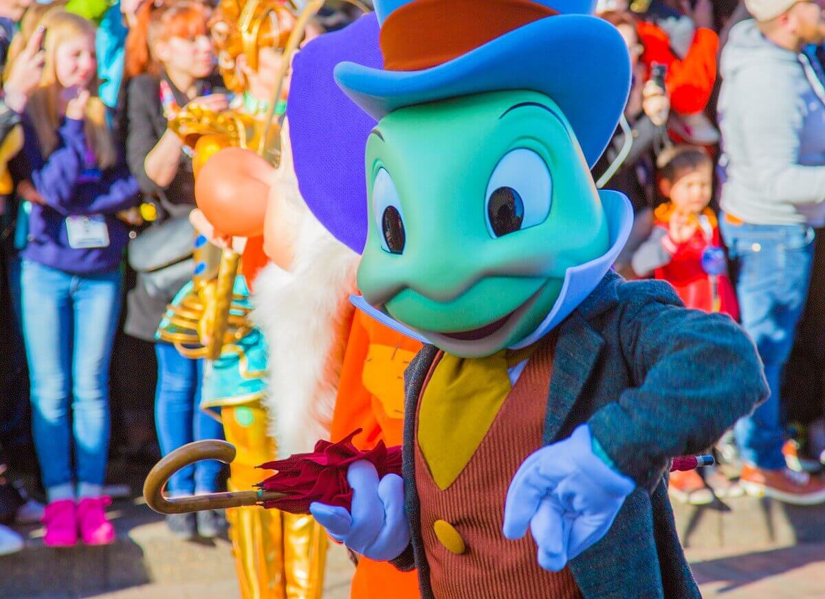 Die Grille Jiminy Cricket aus dem Film Pinocchio hat einen roten Regenschirm unter den Arm geklemmt und tanzt bei der Geburtstagsshow über die Main Street, U.S.A.