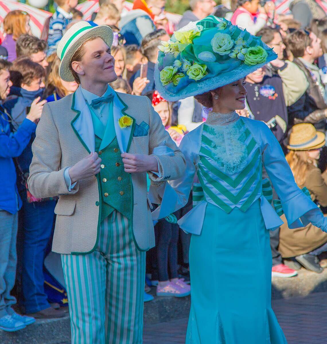 Ein Tanzpaar auf der Main Street, U.S.A. er trägt ein beigefarbenes Sacko und eine grüne Weste, dazu eine beige-grün gestreifte Hose. Sie ein grünes Kleid und einen großen Hut, der mit Blumen besetzt ist