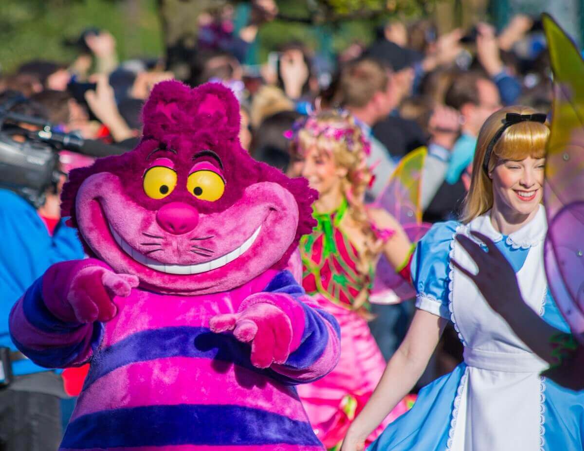 Die Grinsekatze schaut lächelnd in die Kamera, neben ihr läuft Alice im Wunderland