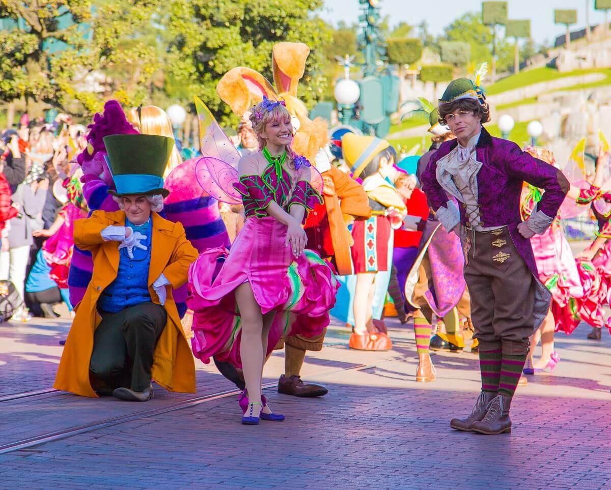Mad Hatter, der verrückte Hutmacher kniet auf der Main Street, U.S.A. neben ihm stehen Tänzer in fantasievollen Kostümen, die an Peter Pan und Feen erinnern