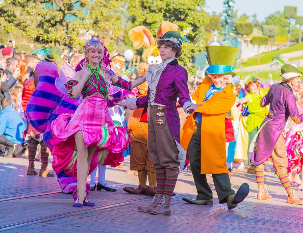 Eine Tänzerin in einem pinken Feenkostüm mit einem Tänzer in einem Kostüm, das an Peter Pan erinnert stehen mit seitlich ausgestreckten Armen auf der Main Street, U.S.A. Hinter ihnen sind der verrückte Hutmacher, der Märzhase und die Grinsekatze zu erkenenn