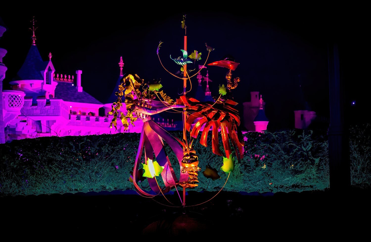 Nächtliche Aufnahme der beleuchteten Skulptur von Aurora und Prinz Phillip in den Gardens of Wonder, im Hintergund die pink angestrahlte Mauer des Disney Schlosses