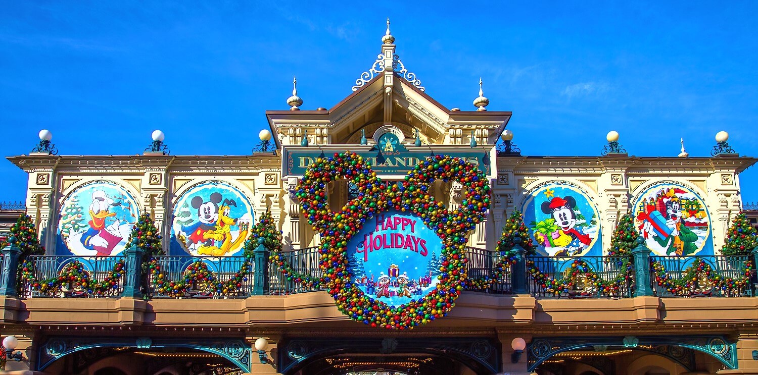 Weihnachtsliche Deko an der Main Street Station: in der Mitte ein Weihnachtskrank in der Form eines Mickey Mouse Kopfes und einem Schild mit Chip & Dale auf dem 'Happy Holidays' steht, rechts und links Tafeln mit weihnachtlichen Motiven mit Donald Duck, Mickey Mouse, Pluto, Minnie Mouse und Goofy