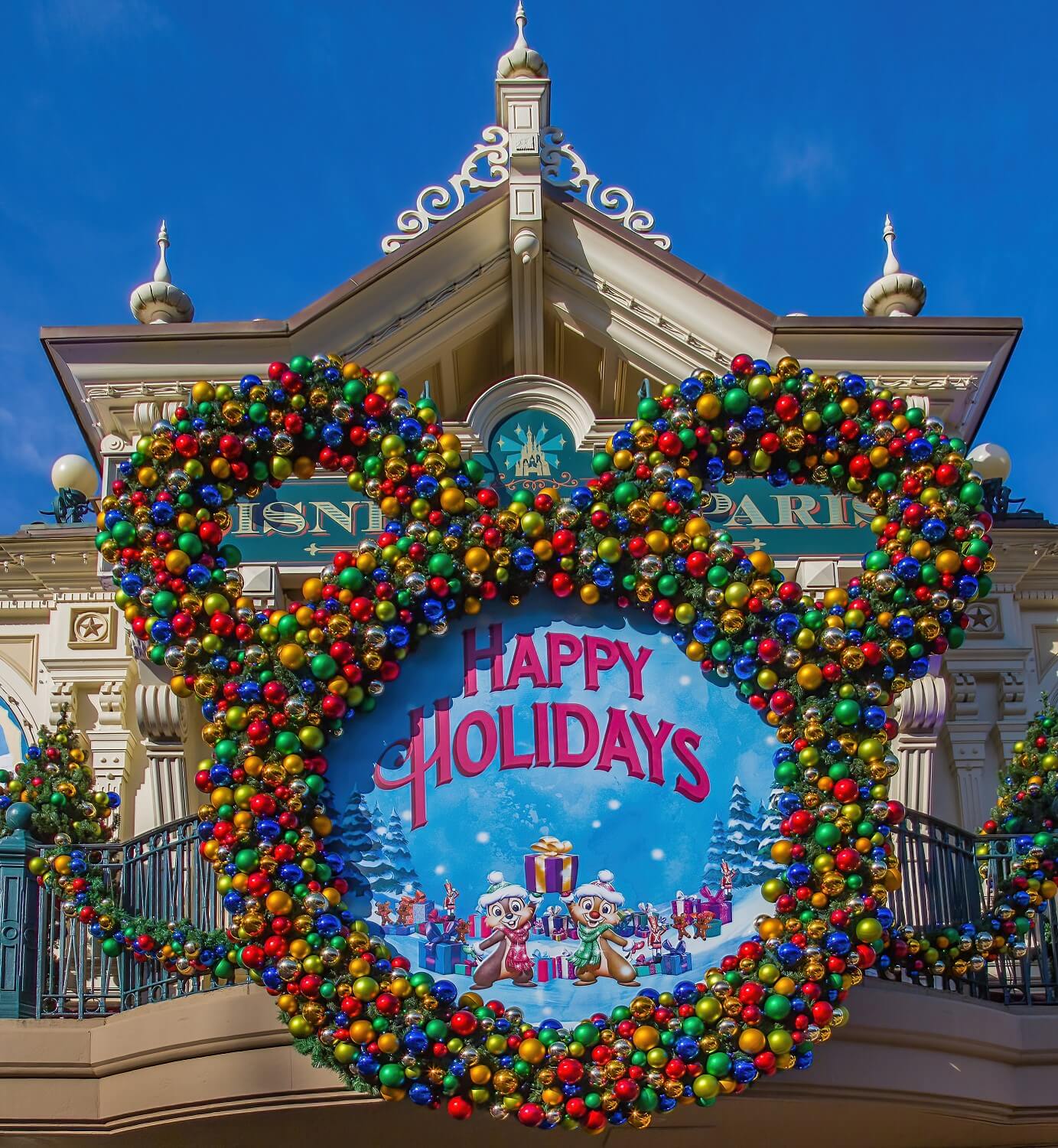 Weihnachtskranz in Form eines Mickey-Kopfes mit roten, blauten, grünen und goldenen Kugeln und einem 'Happy Holidays' Schild mit Chip & Chap in der Mitte