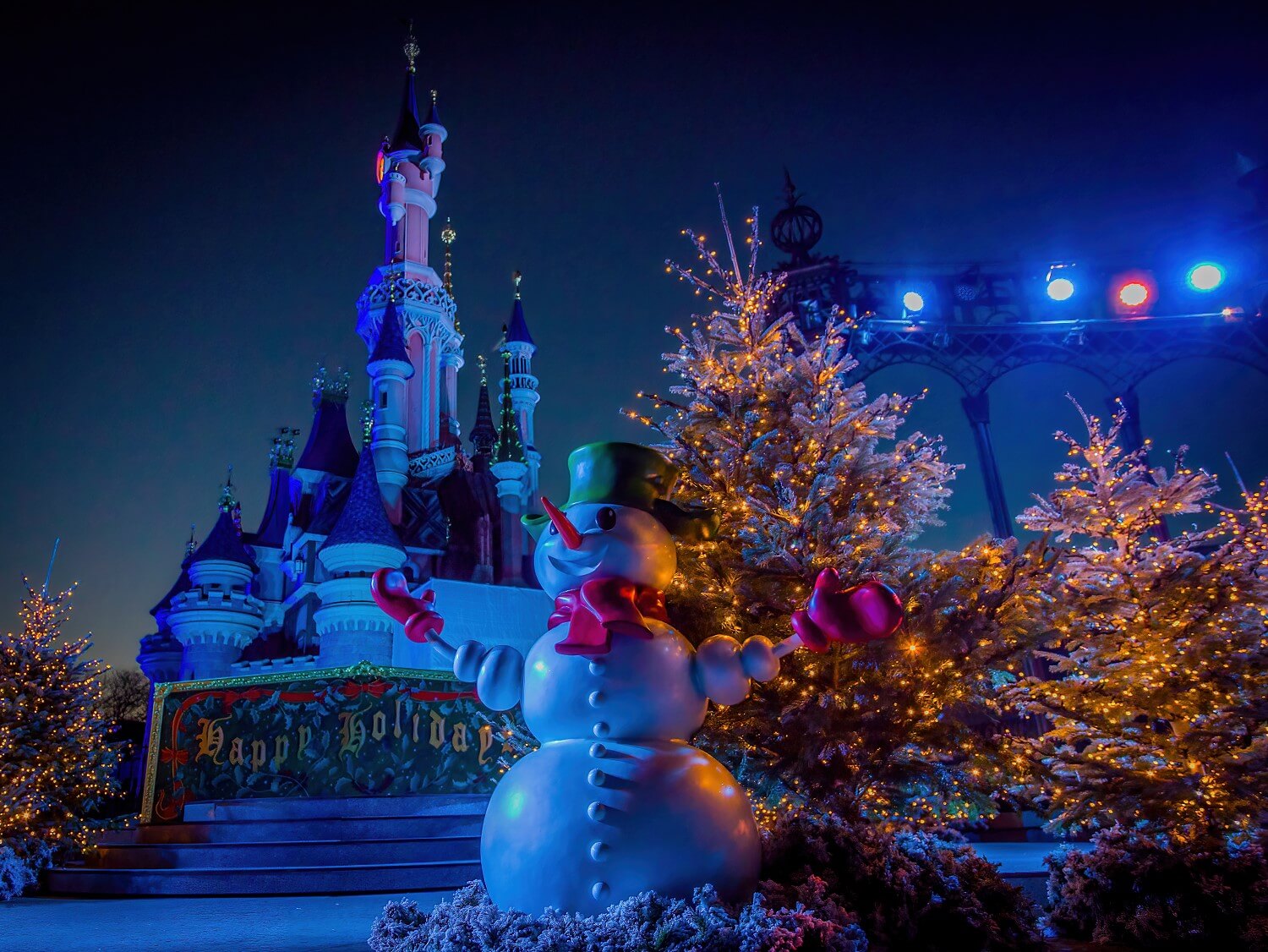 Schneemann mit grüner Mütze und roten Handschuhen auf der Castle Stage. Rechts und links hinter ihm Bäume mit hunderten kleiner Lichter, im Hintergrund ein Schild 'Happy Holidays' und das nächstlich angestrahlte Dornröschenschloss