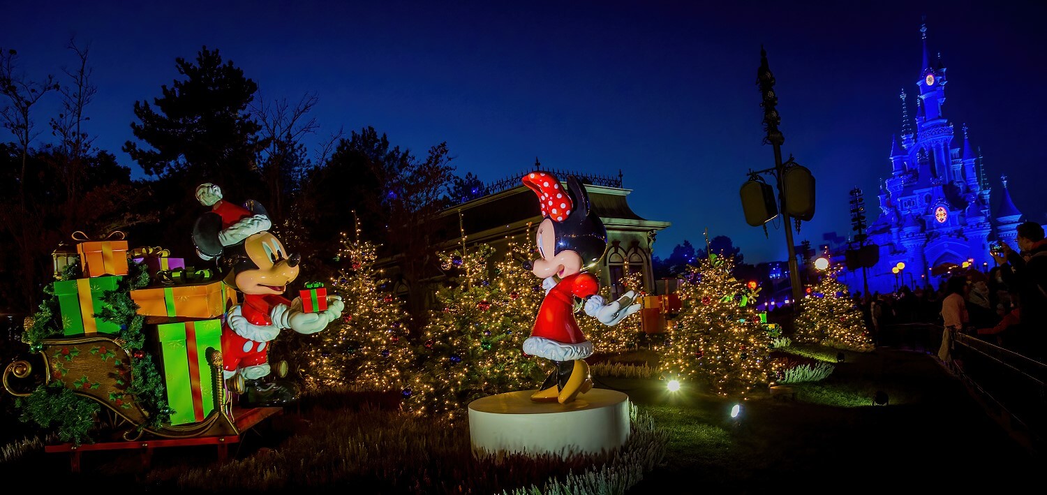 Mickey und Minnie in einem weihnachtlich beleuchteten Wäldchen bei Nacht. Mickey bringt Minnie mit einem Schlitten viele Geschenke, recht im Hintergrund das Schloss