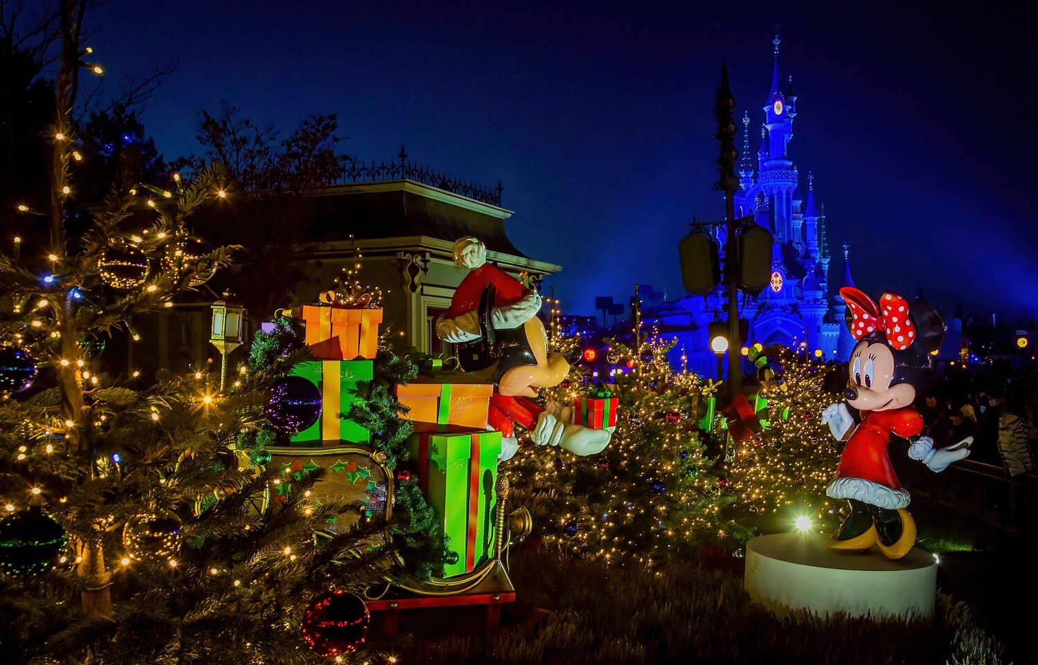 Mickey gibt der Minnie ein kleines Geschenk, viele weitere liegen auf einem Schlitten hinter ihm. Die beiden sind von kleinen Weihnachtsbäumen mit hunderten kleiner Lichter umgeben