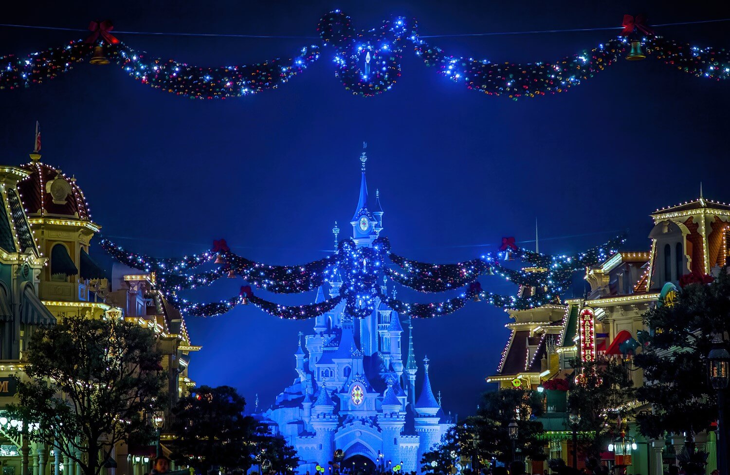 Die Main Street USA bei Nacht, überall leuchten die Lichter und der Weihnachtschmuck, im Hintergrund ist das Schloss des Disneyland Park blau angestrahlt
