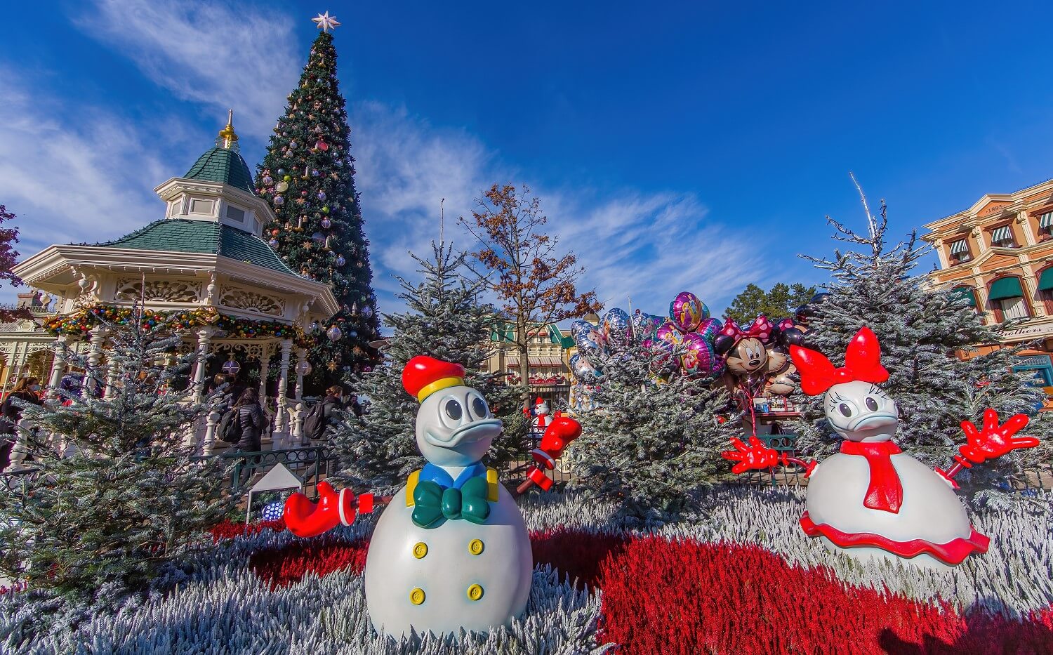 Weitwinkelfoto mit Donald Duck und Daisy Duck als Schneemännern am Town Square mit Gazebo und Weihnachtsbaum links dahinter und dem Geschäft der Bixby Brothers rechts im Hintergrund