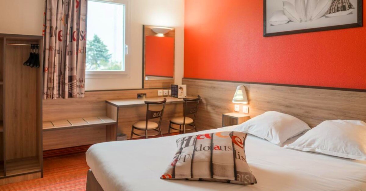 Hotelzimmer mit einer rot gestrichenen Wand und einem Doppelbett, vor dem Fenster steht ein kleiner Tisch mit zwei Stühlen